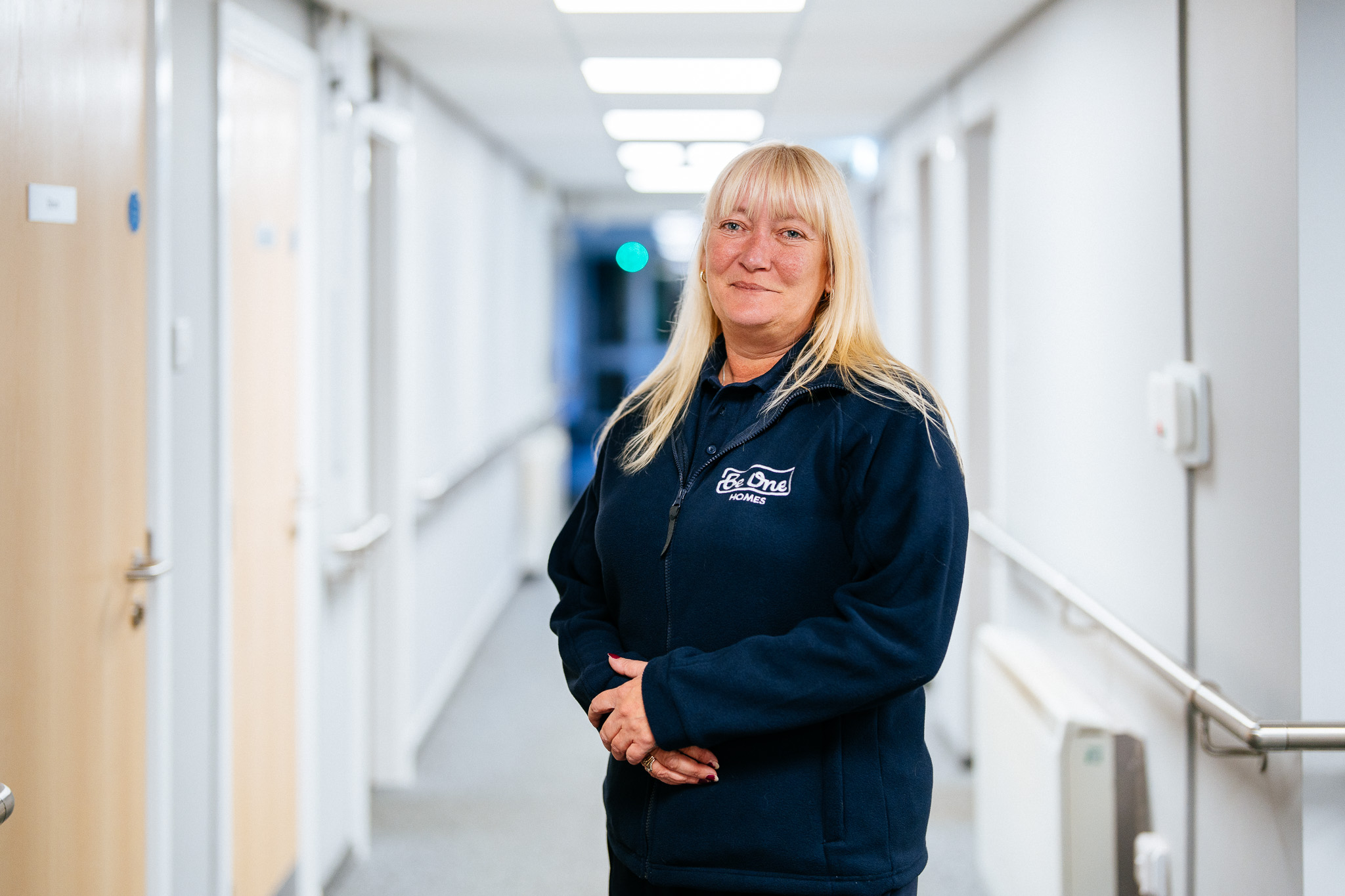 A female colleague from the Careline Team standing in a well-lit corridor, wearing a blue Be One Homes jumper and holding her hands together in front of her.