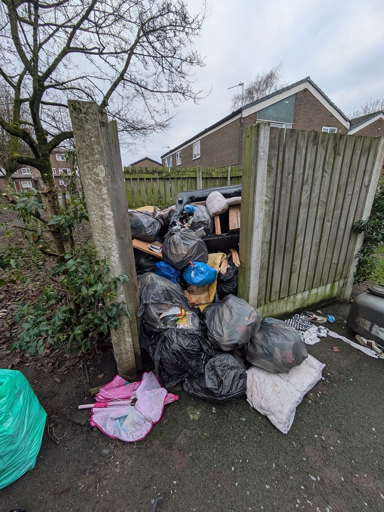 A large pile of fly‑tipped rubbish blocking an entrance beside a wooden fence. The pile includes black and blue bin bags, an old sofa, children’s toys, shoes, an umbrella, and loose litter scattered on the ground.