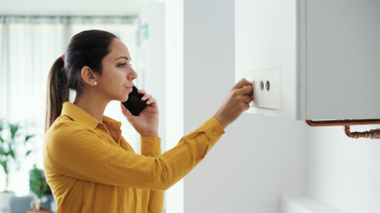 A lady holding a mobile phone to her ear whilst adjusting a dial on her boiler. She is wearing a yellow shirt and is stood in a room with white walls.