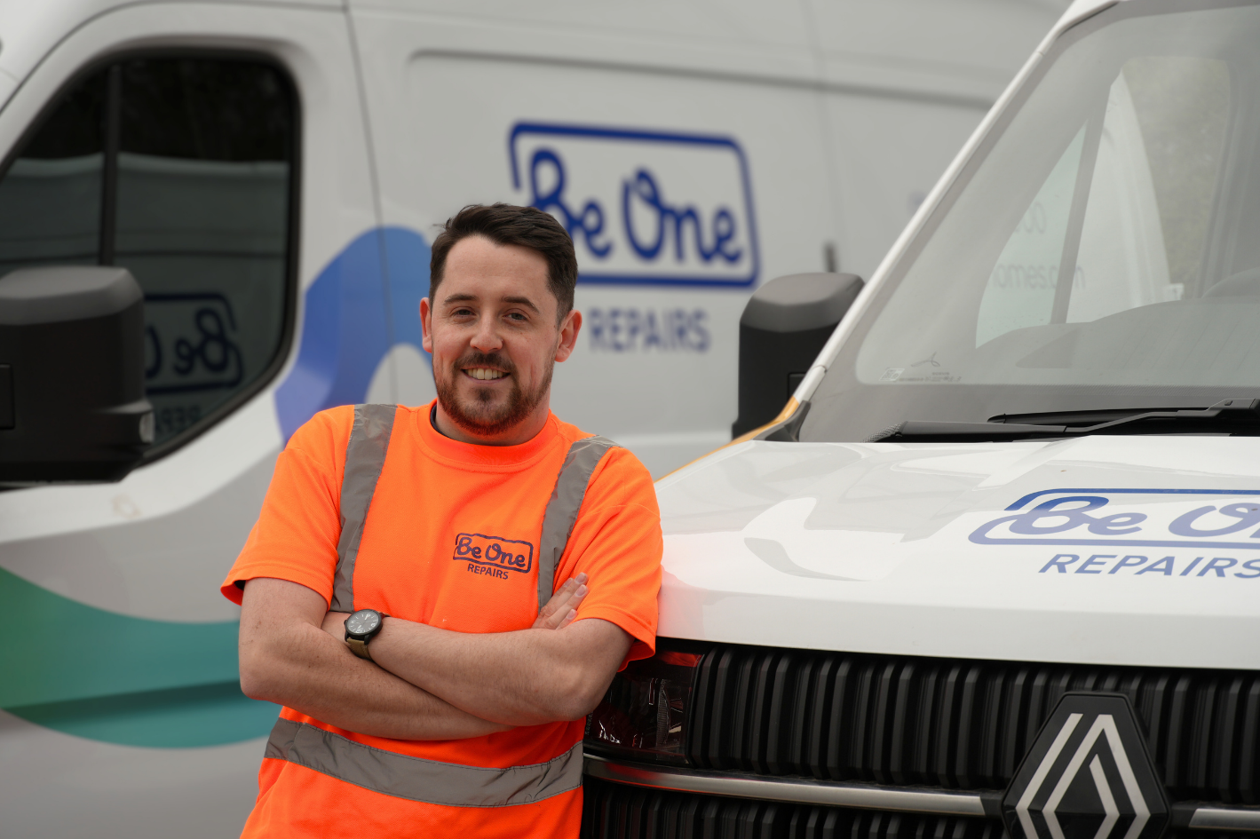 A male Joiner wearing an orange Be One Homes hi-visibility jacket. He is stood next to a Be One Homes van with his arms folded.