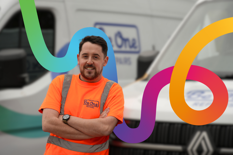A male Joiner wearing an orange Be One Homes hi-visibility jacket. He is stood next to a Be One Homes van with his arms folded. This version of the image includes the multicoloured Be One Homes brand thread.