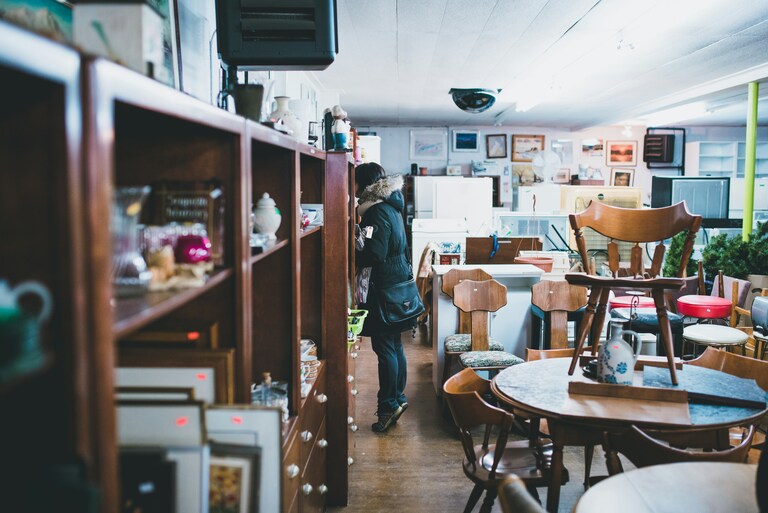 The interior of a second-hand furniture shop. A woman is looking at some of the wooden shelves to the left of the image. There are dining tables and chairs on the right.