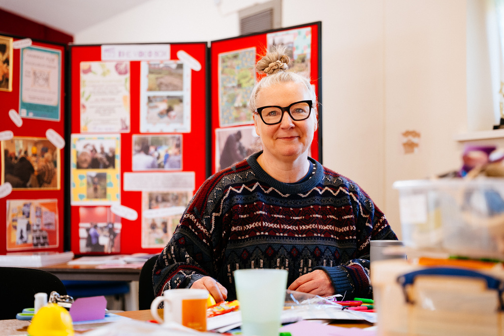 A person sitting at a table with craft materials, looking toward the camera. Behind the person is a bright red display board filled with photos and information about community projects.
