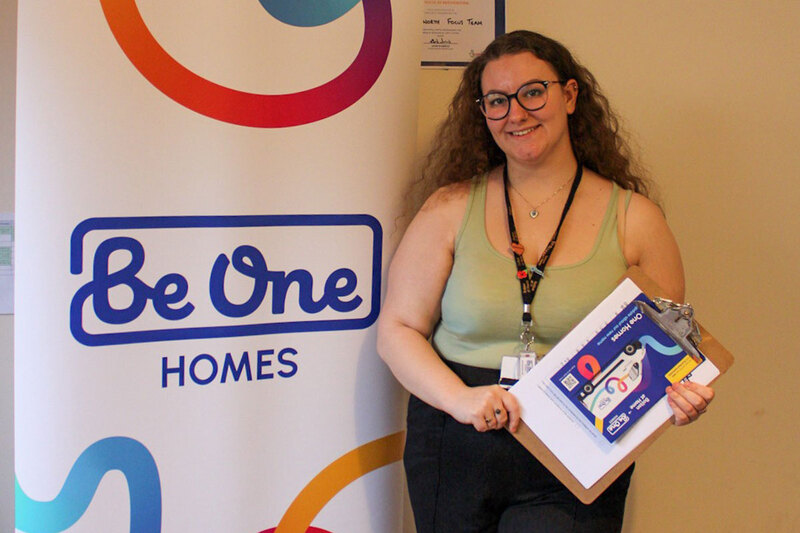 A colleague wearing a lanyard stands next to a “Be One Homes” branded banner, holding a clipboard with printed materials.