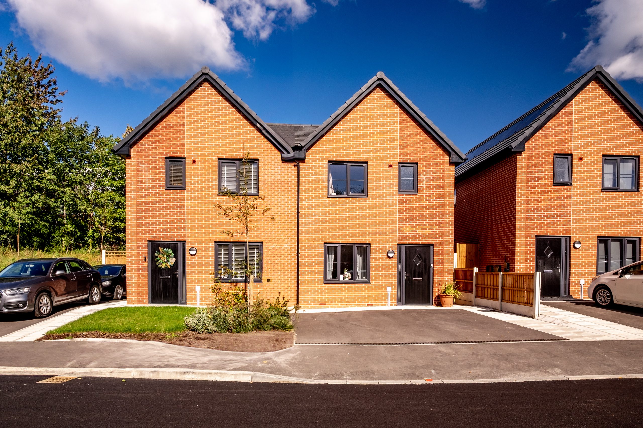 A pair of modern semi-detached Be One Homes properties featuring red brick exteriors with dark grey pitched roofs and black front doors. Each home has large windows, a small front garden area with newly planted greenery, and a driveway with parked cars. The houses sit along a clean, newly paved residential street with a bright blue sky and scattered clouds above, and green trees visible in the background.