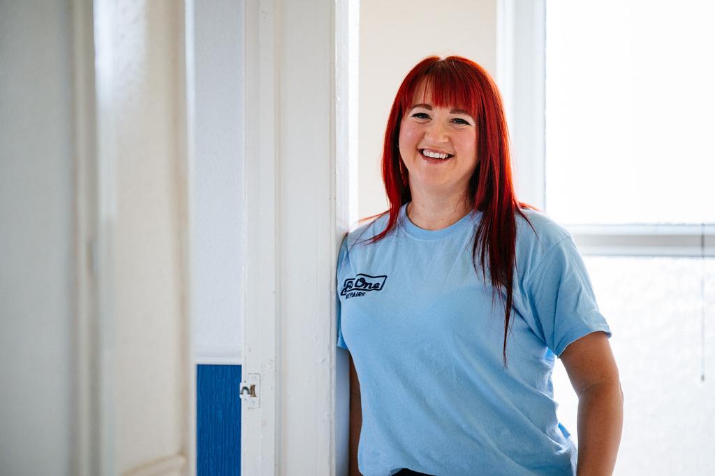 A female Be One Homes colleague wearing a light blue Be One Homes-branded jumper. She is leaning against a door frame and is smiling at the camera.