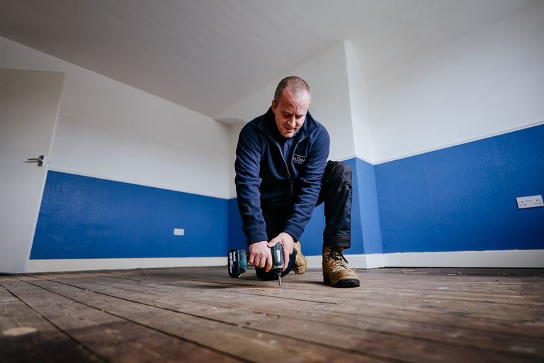 Image of a male Be One Repairs operative using a drill to fix a floorboard.