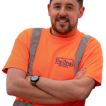 A male Joiner wearing an orange Be One Homes hi-visibility jacket. He is stood next to a Be One Homes van with his arms folded.
