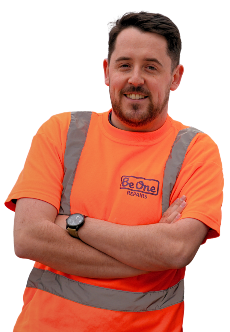 A male Joiner wearing an orange Be One Homes hi-visibility jacket. He is stood next to a Be One Homes van with his arms folded.