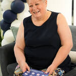 Image of a lady sat and smiling at something whilst holding a Be One Homes leaflet in her hands. Blue and white balloons are behind her.