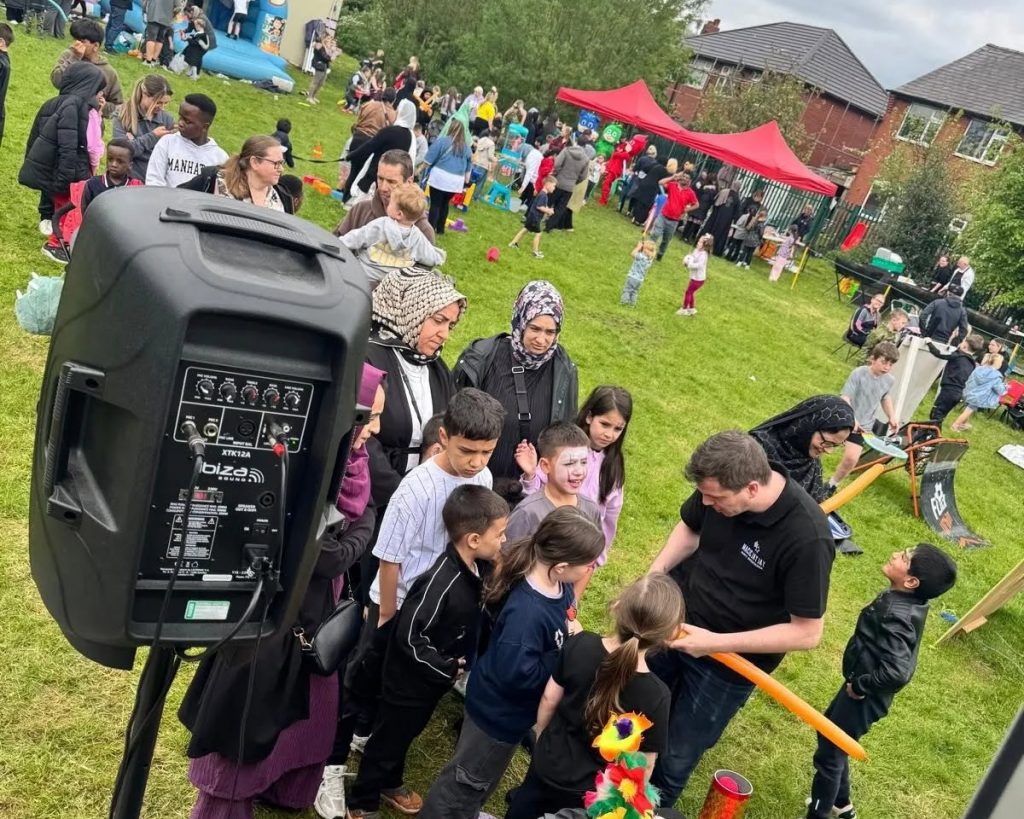 A community event in one of our neighbourhoods. There are many people dispersed around a field. A speaker system is visible in the foreground, with gazebos visible in the distance.