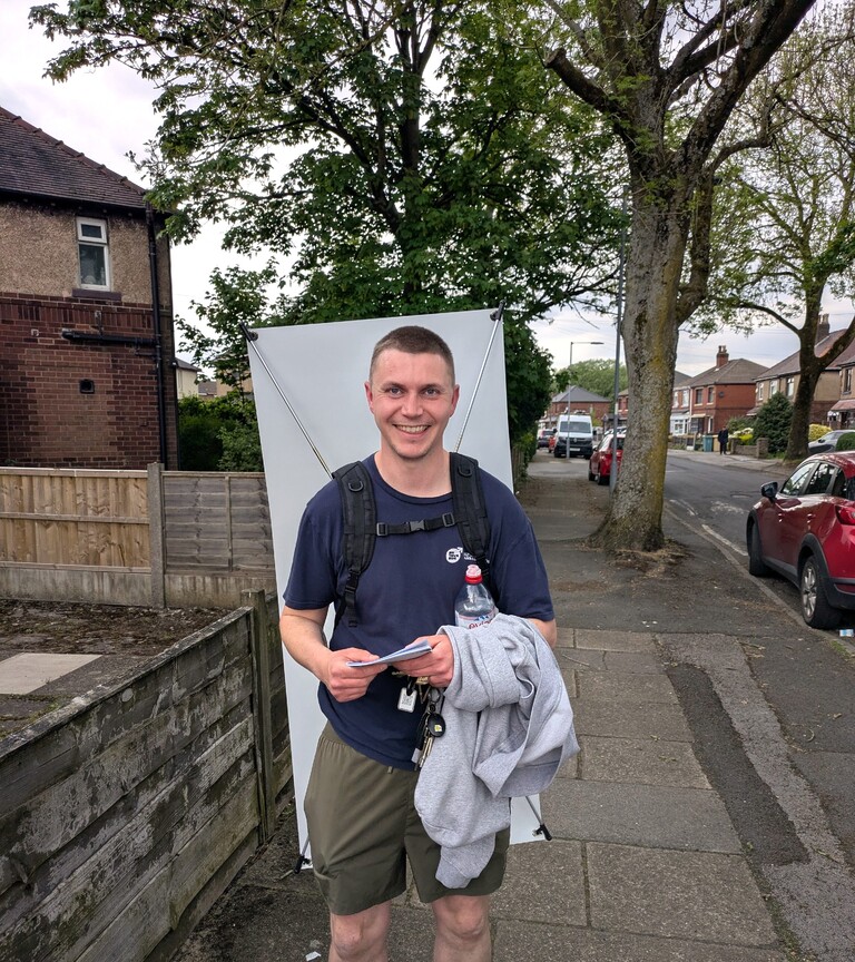 Image of Dan Livesey, our Bolton Manbassadors Project Officer. Dan is smiling, looking at the camera, and is wearing a sign promoting the Bolton Manbassadors programme on his back.